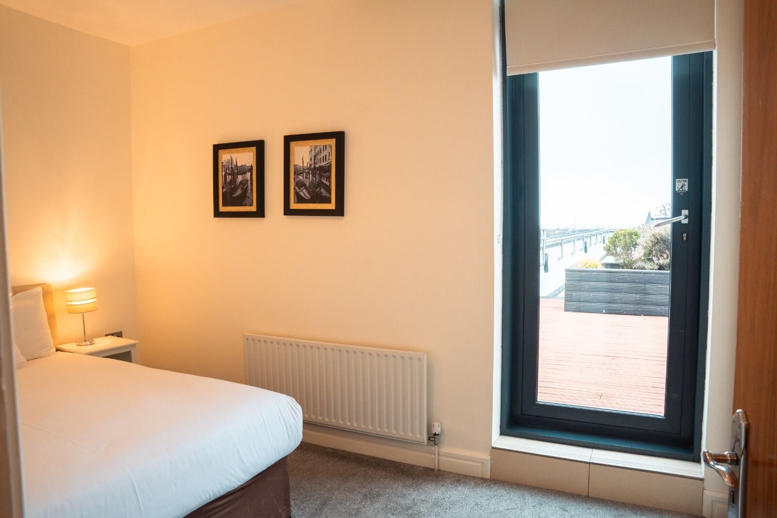 A hotel room with a bed, a bedside lamp, a radiator, and a window with a view of a building and a brick wall.