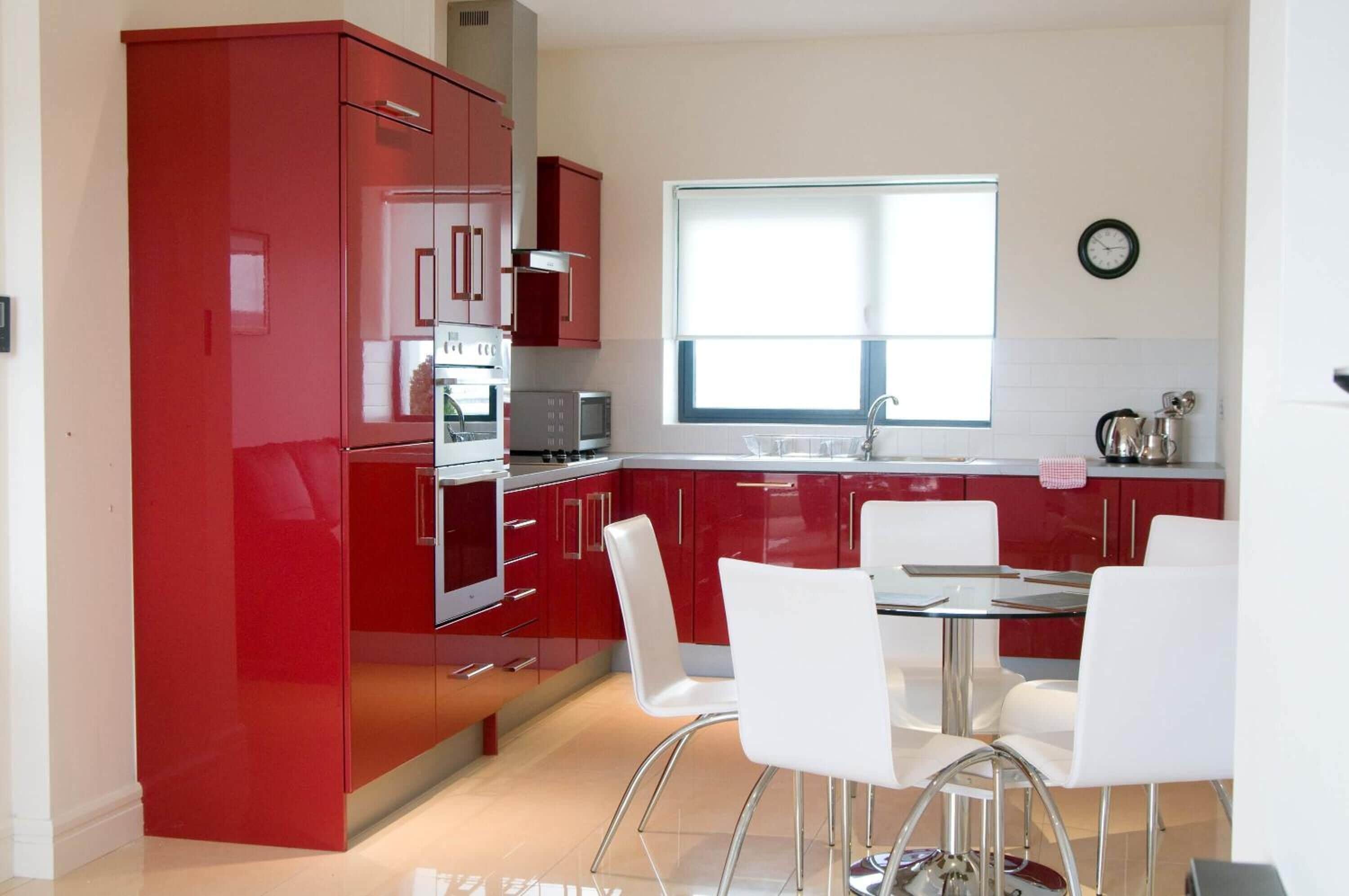 A modern kitchen with red cabinets, a stainless steel oven, and a dining area with white chairs.