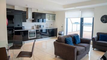 A modern kitchen with a microwave, oven, and cabinets. There is a dining area with a glass table and chairs. A sofa and armchair are in the living area, and a large window offers a view of the outside.