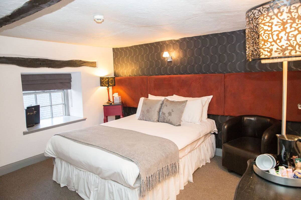 A hotel room with a large bed, a red headboard, a window with blinds, a bedside table with a lamp, and a black leather armchair.