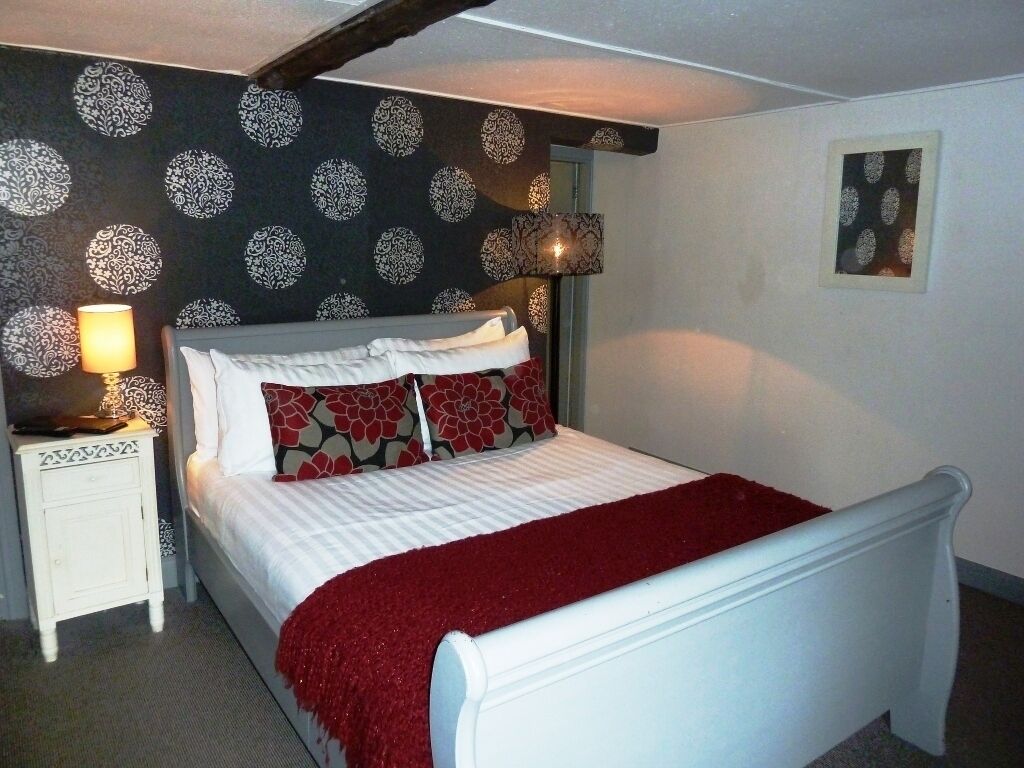 Une chambre à coucher avec un lit, une table de chevet blanche, un papier peint à motifs et un plaid rouge.