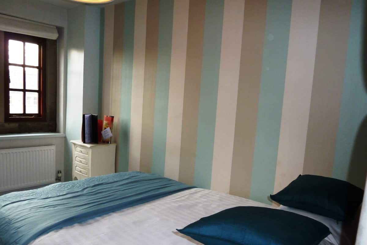 A bedroom with a striped wall, a window, a radiator, a dresser, and a bed with pillows.