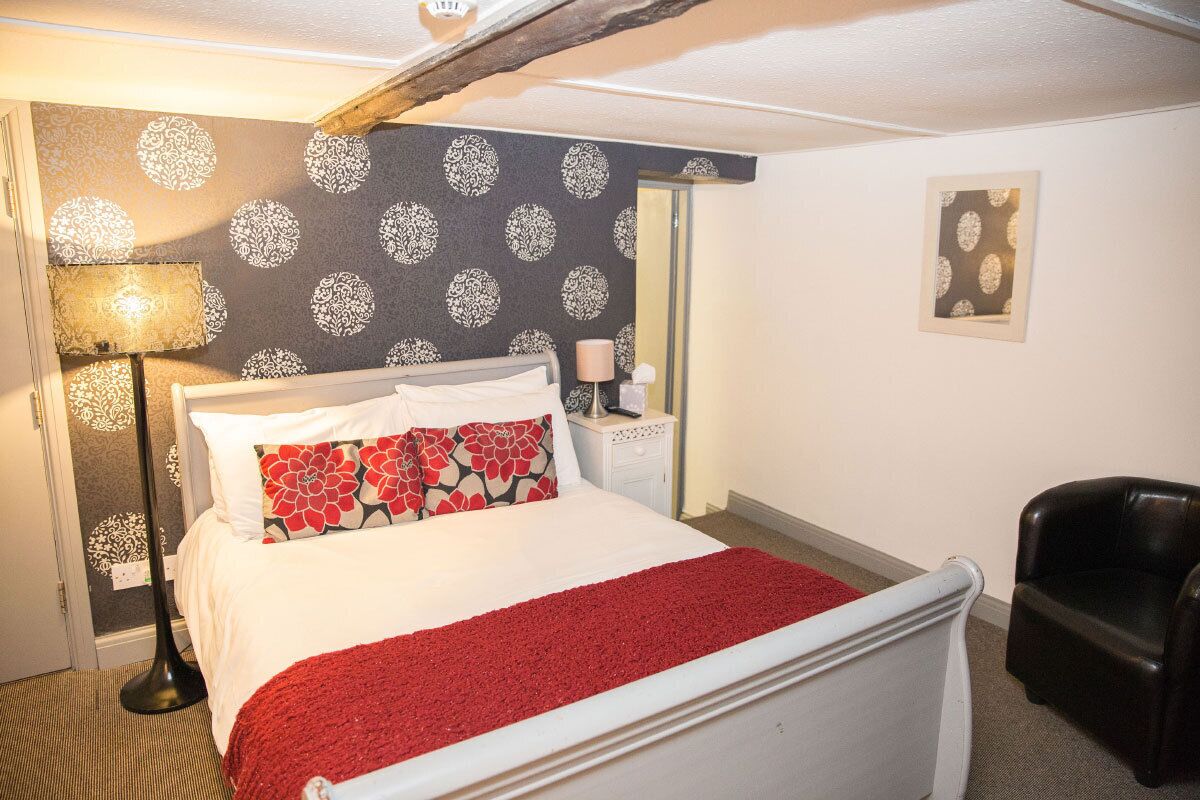 A bedroom with a bed, a red throw blanket, a nightstand, a lamp, a chair, and a patterned wallpaper.