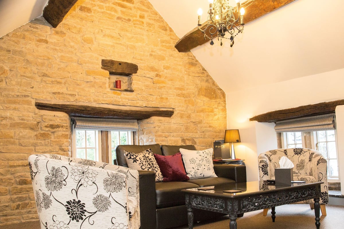 A cozy living room with a stone wall, a black sofa, floral-patterned armchairs, and a wooden coffee table.