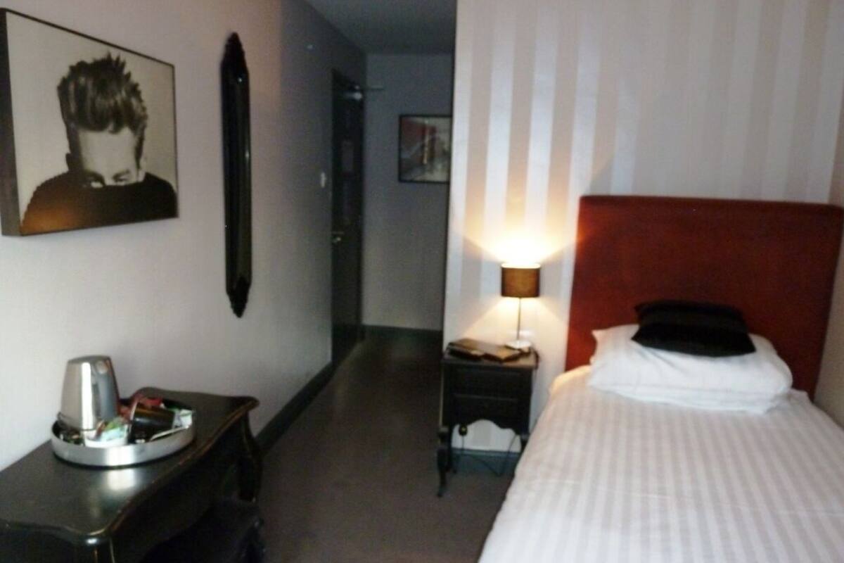 A hotel room with a bed, a bedside table with a lamp, a picture on the wall, and a small table with a coffee maker.