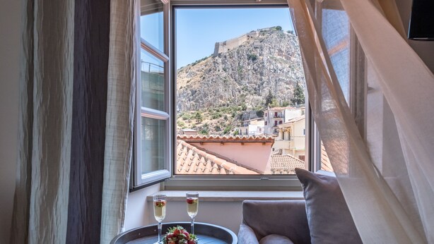A room with a view of a mountainous landscape through a window, a round table with a bowl of fruit and two glasses of wine, and a grey sofa. - Grand Sarai Nafplio (Nafplio)