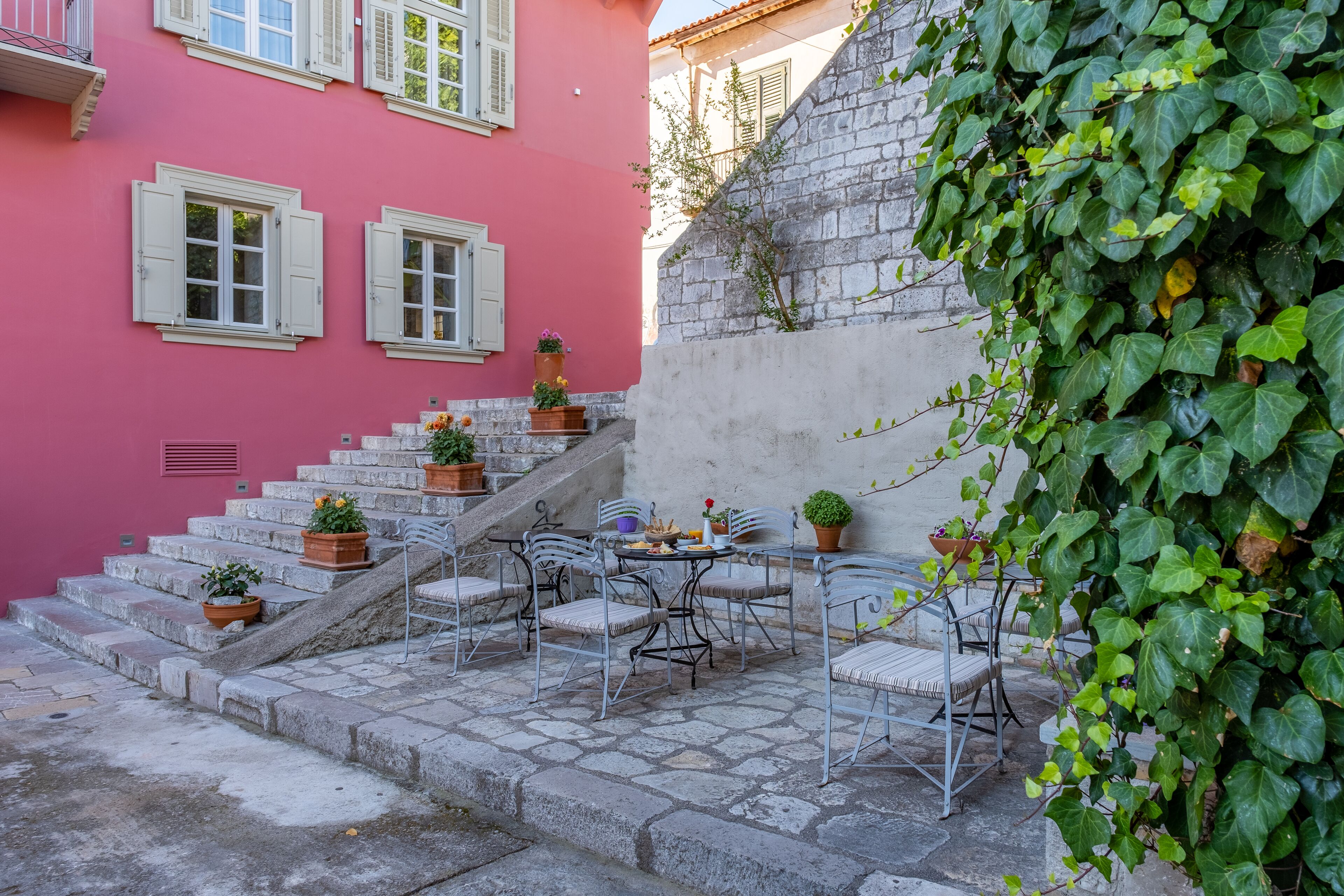 Un escalier en pierre mène à un bâtiment rose aux volets blancs. On y trouve des tables et des chaises à l’extérieur, ainsi que des plantes en pot.