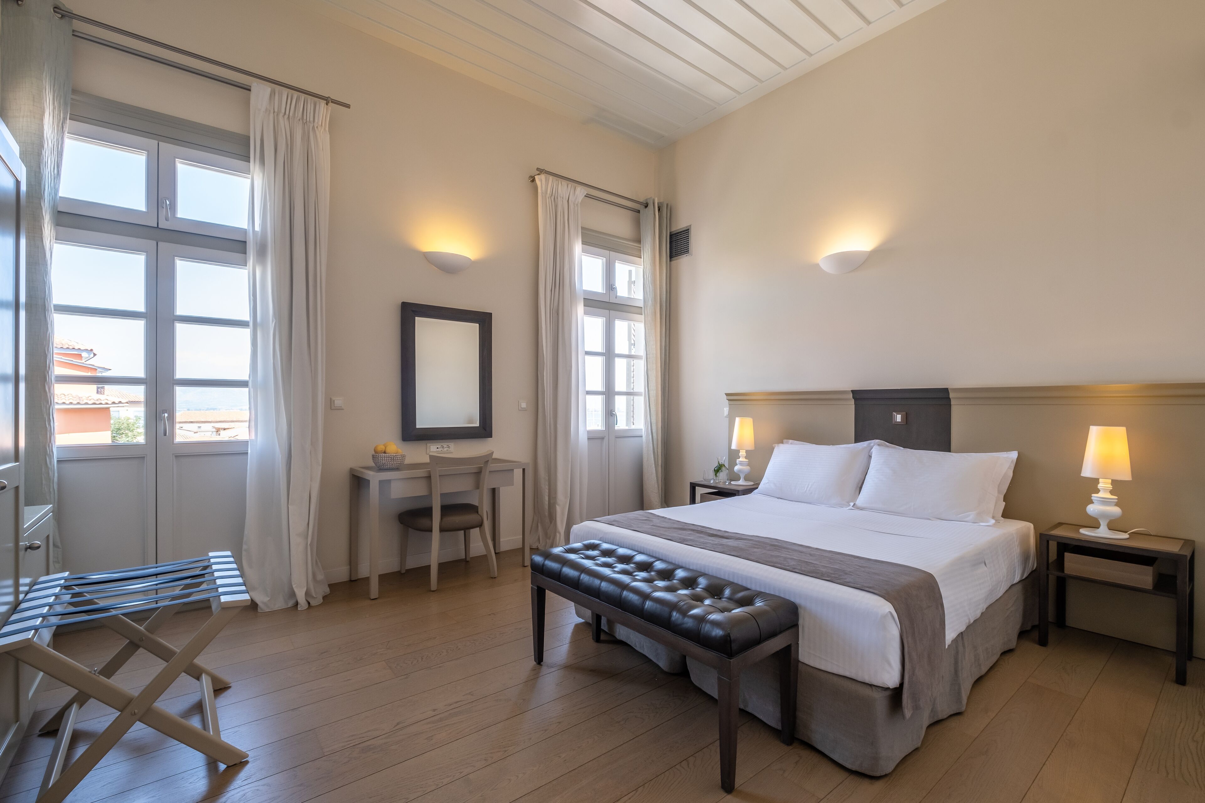 A hotel room with a bed, a desk, a chair, a bench, and a window with curtains.