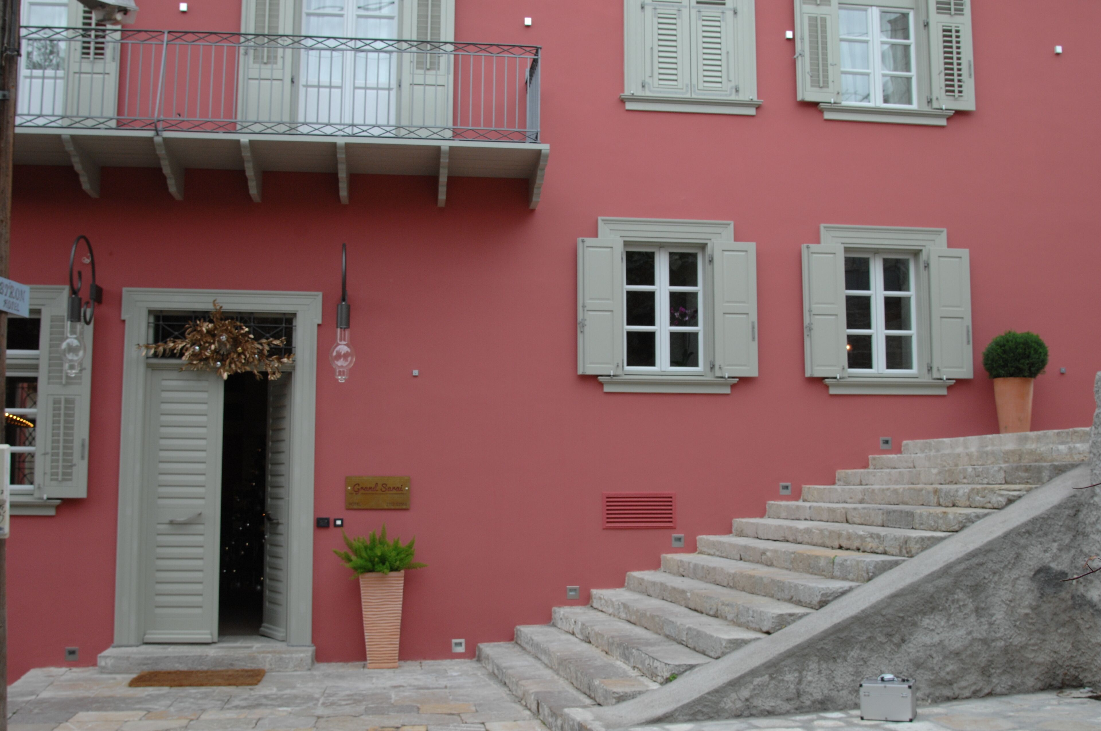 A building with a pink facade, white shutters, and a stone staircase leading to a door with a hanging wreath.