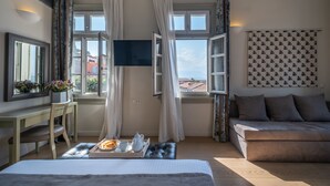 A hotel room with a bed, a sofa, a TV, a desk with a chair, and a window with curtains. - Grand Sarai Nafplio (Nafplio)