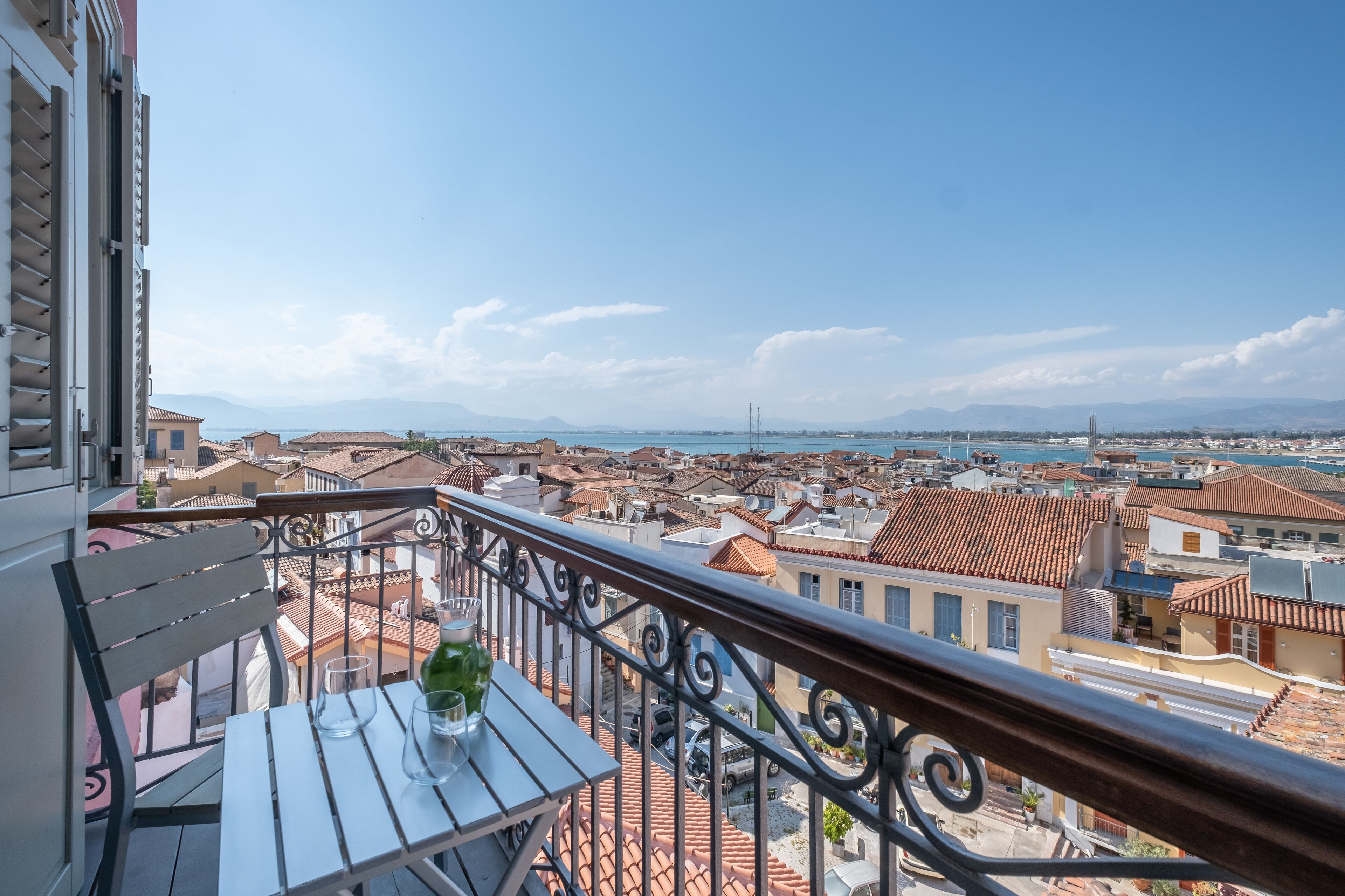 Un balcon avec une table et des chaises, donnant sur un paysage urbain avec des toits recouverts de tuiles rouges et une vue lointaine sur la mer.