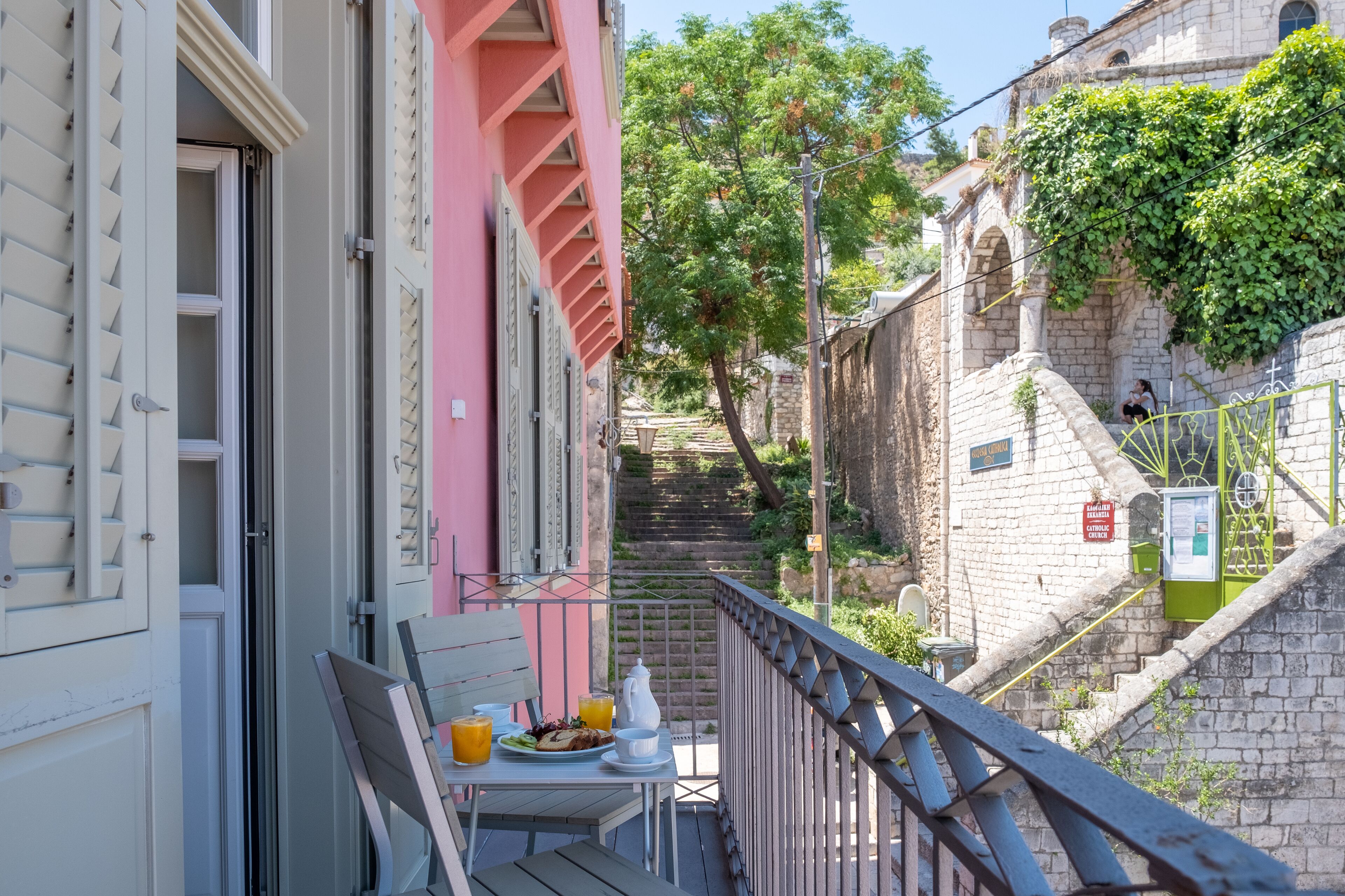 Ein Balkon mit Frühstückstisch, der auf eine enge Straße mit einer Treppe zu einem Gebäude mit einem Schild blickt.