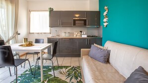 Una cocina moderna con armarios oscuros, una encimera blanca y una pared de acento color turquesa. Cuenta con una zona de comedor con una mesa y sillas blancas, y un sofá color beige con cojines grises.