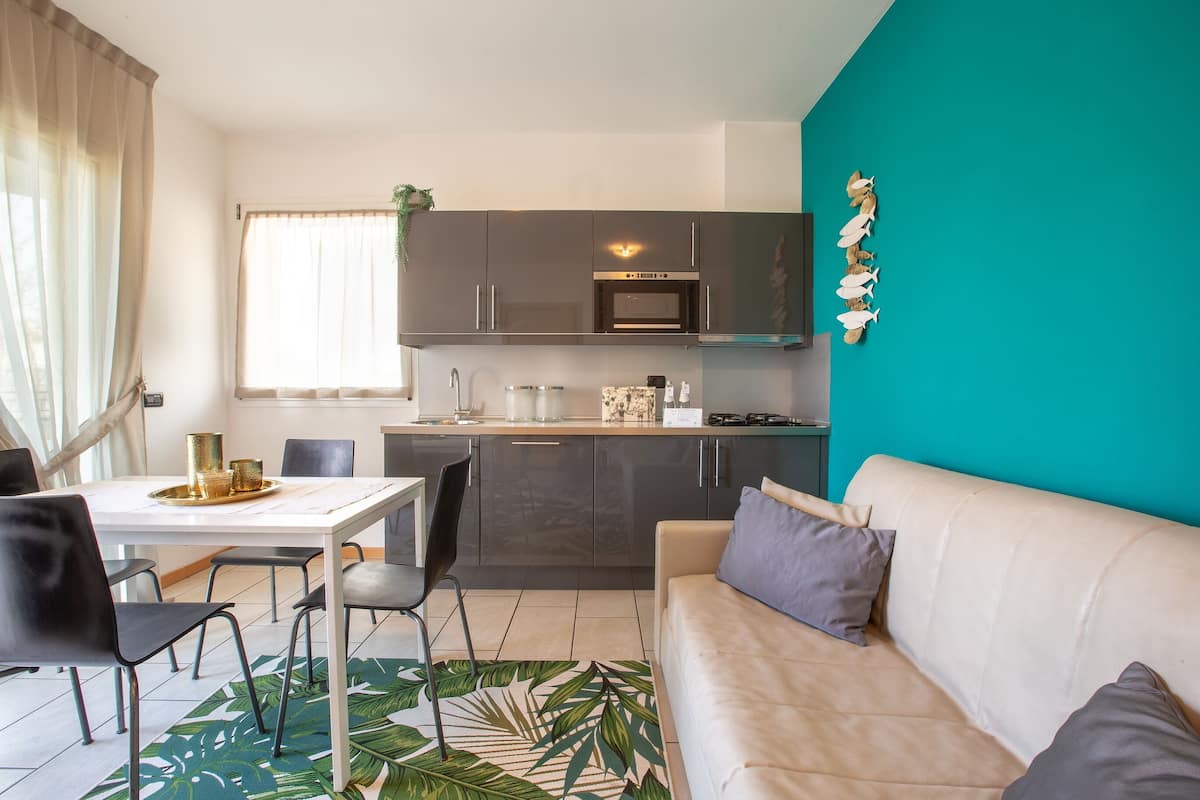 A modern kitchen with dark cabinets, a white countertop, and a teal accent wall. There is a dining area with a white table and chairs, and a beige sofa with gray cushions.