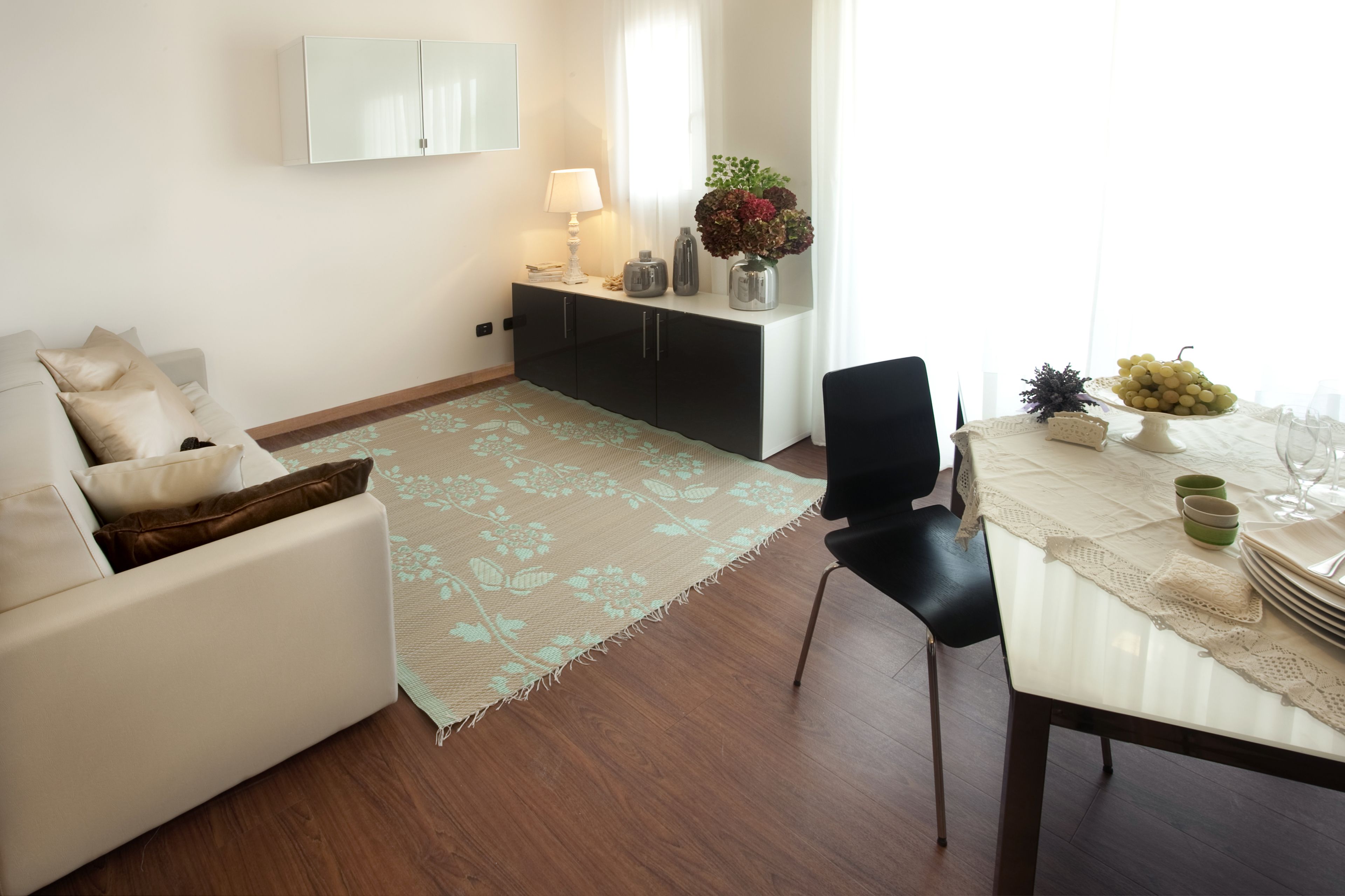 A modern living room with a sofa, a dining table set with a vase of flowers, and a cabinet with a lamp.