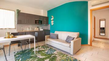 A compact living space with a kitchenette, a white sofa, a dining table with chairs, and a tropical leaf-patterned rug.