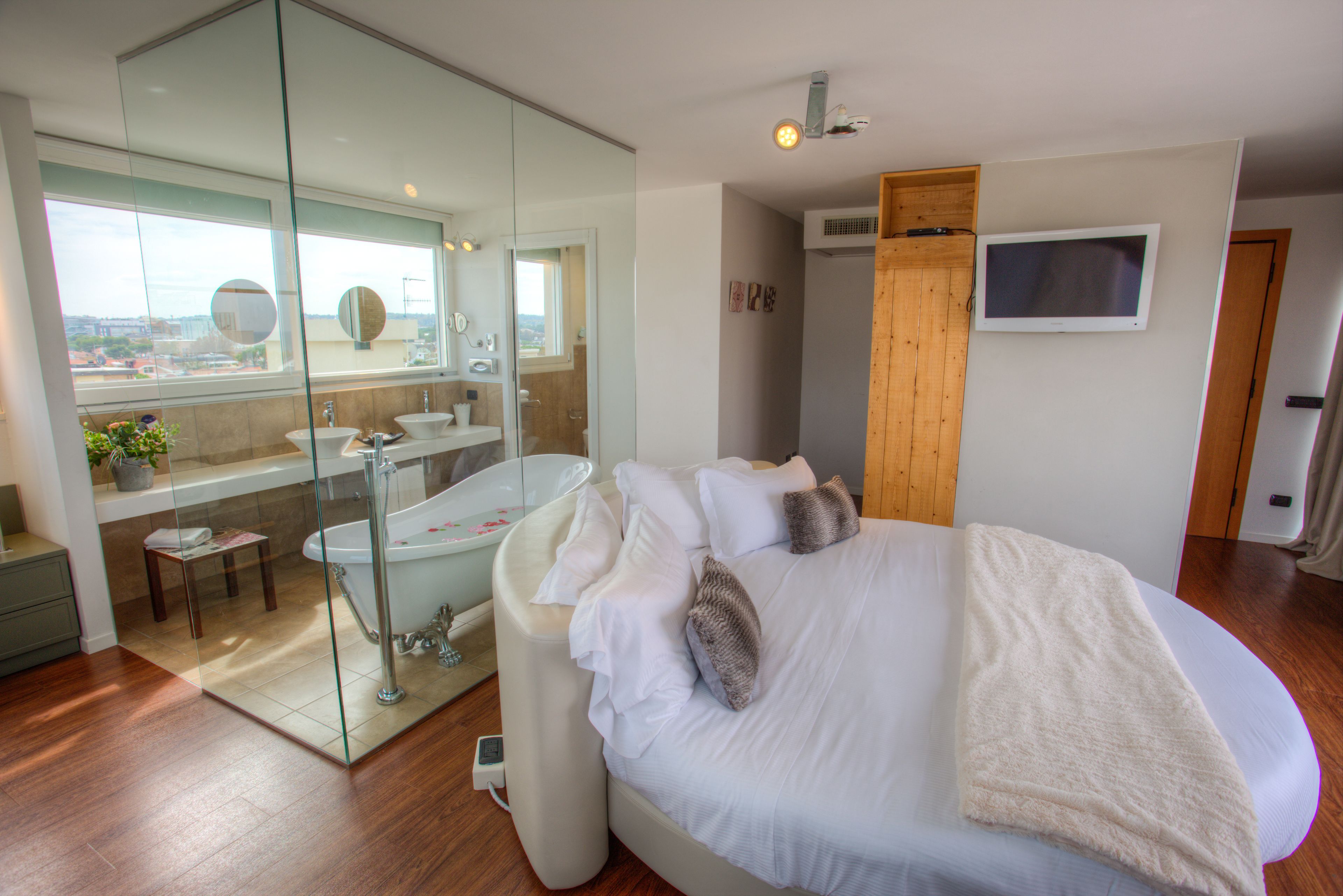 A modern bedroom with a large bed, a glass-enclosed bathroom, a TV mounted on the wall, and a view of the city through the window.