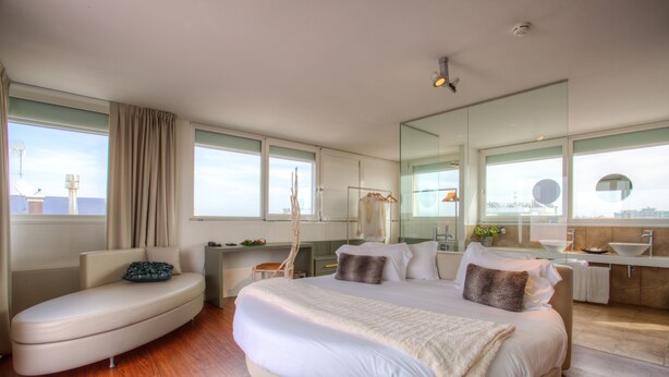 Una habitación de hotel moderna con una cama grande, un área de descanso, un escritorio y un baño con un espejo amplio y lavamanos.