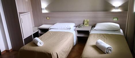 Una habitación de hotel con dos camas individuales, cada una con una colcha color beige y una toalla blanca enrollada. Hay mesitas de noche con un teléfono y un cartel, y un armario con espejo.