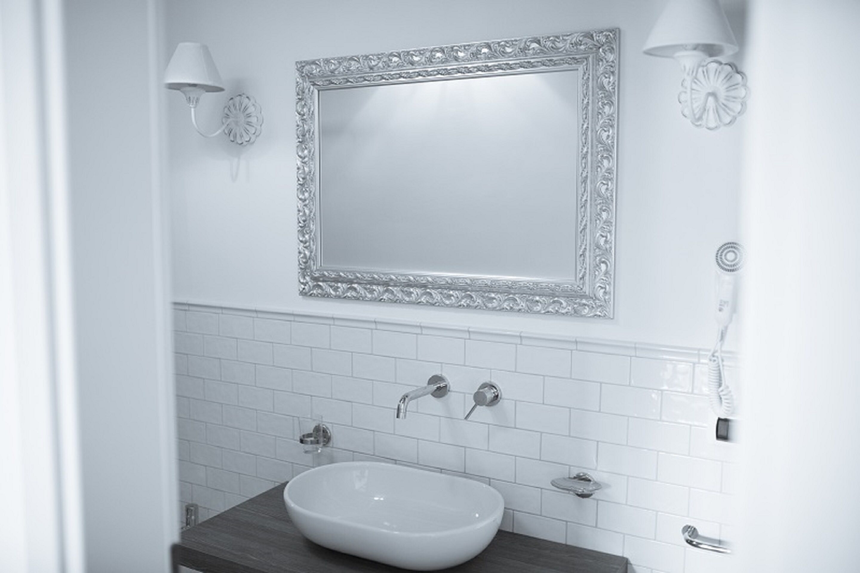 Un bagno con un lavandino bianco, due rubinetti, uno specchio con cornice decorata e un portalampada a parete.