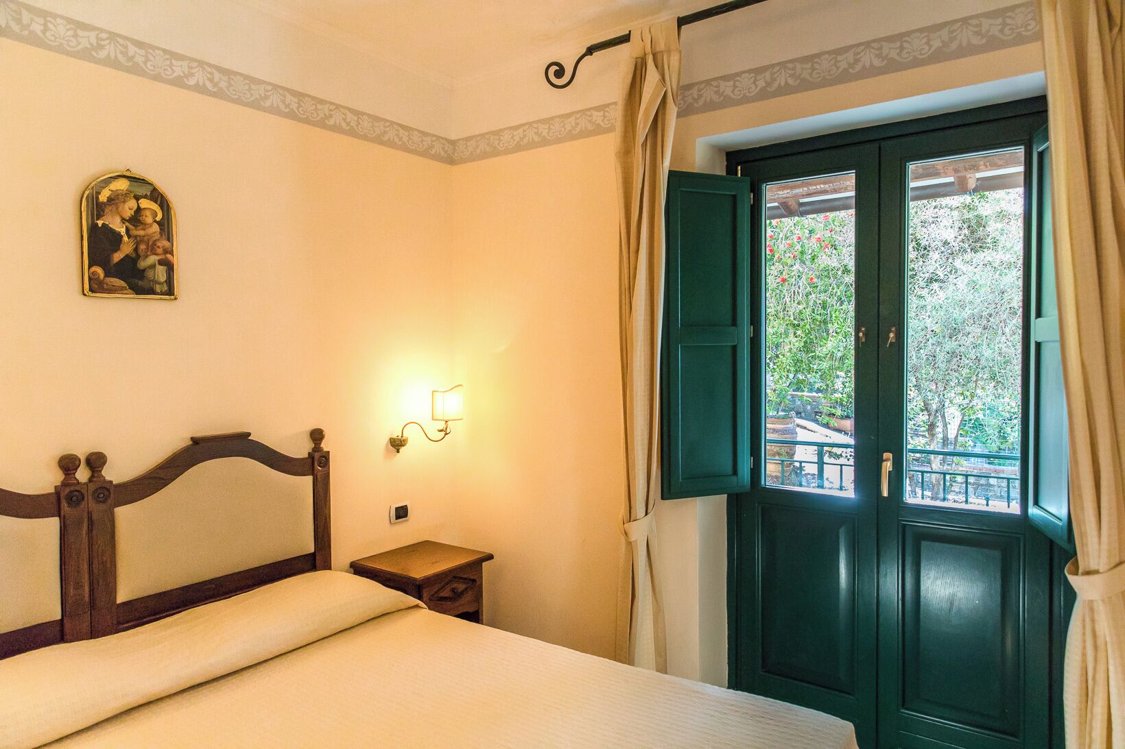 A bedroom with a bed, a nightstand, a wall-mounted light, a window with green shutters, and a decorative wall piece.