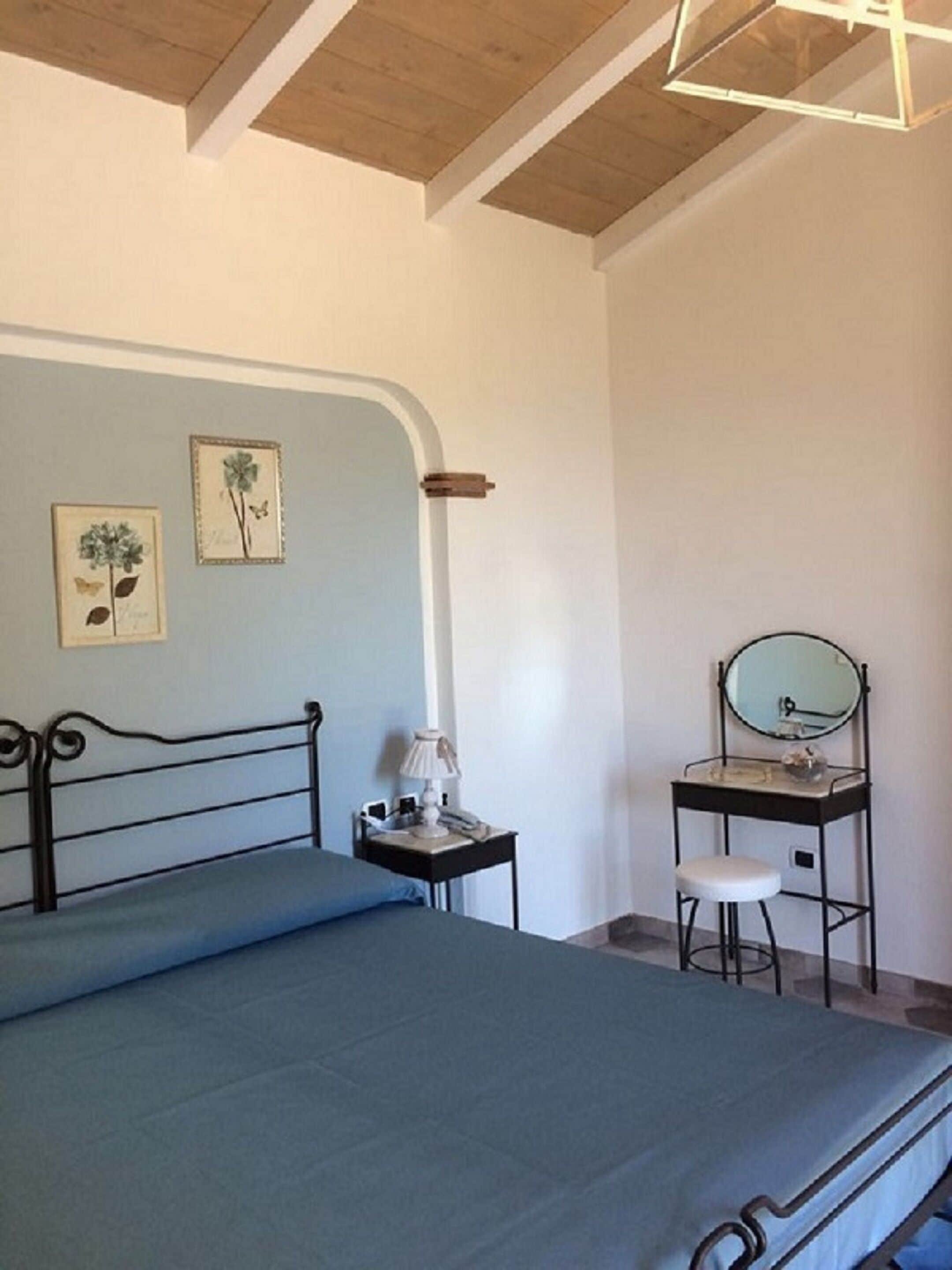Quarto com cama, mesa de cabeceira com luminária, um banquinho pequeno, espelho e duas gravuras botânicas emolduradas na parede.
