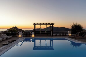 Una piscina en la azotea con una pérgola, sillas de descanso y vistas a la puesta de sol sobre las montañas.