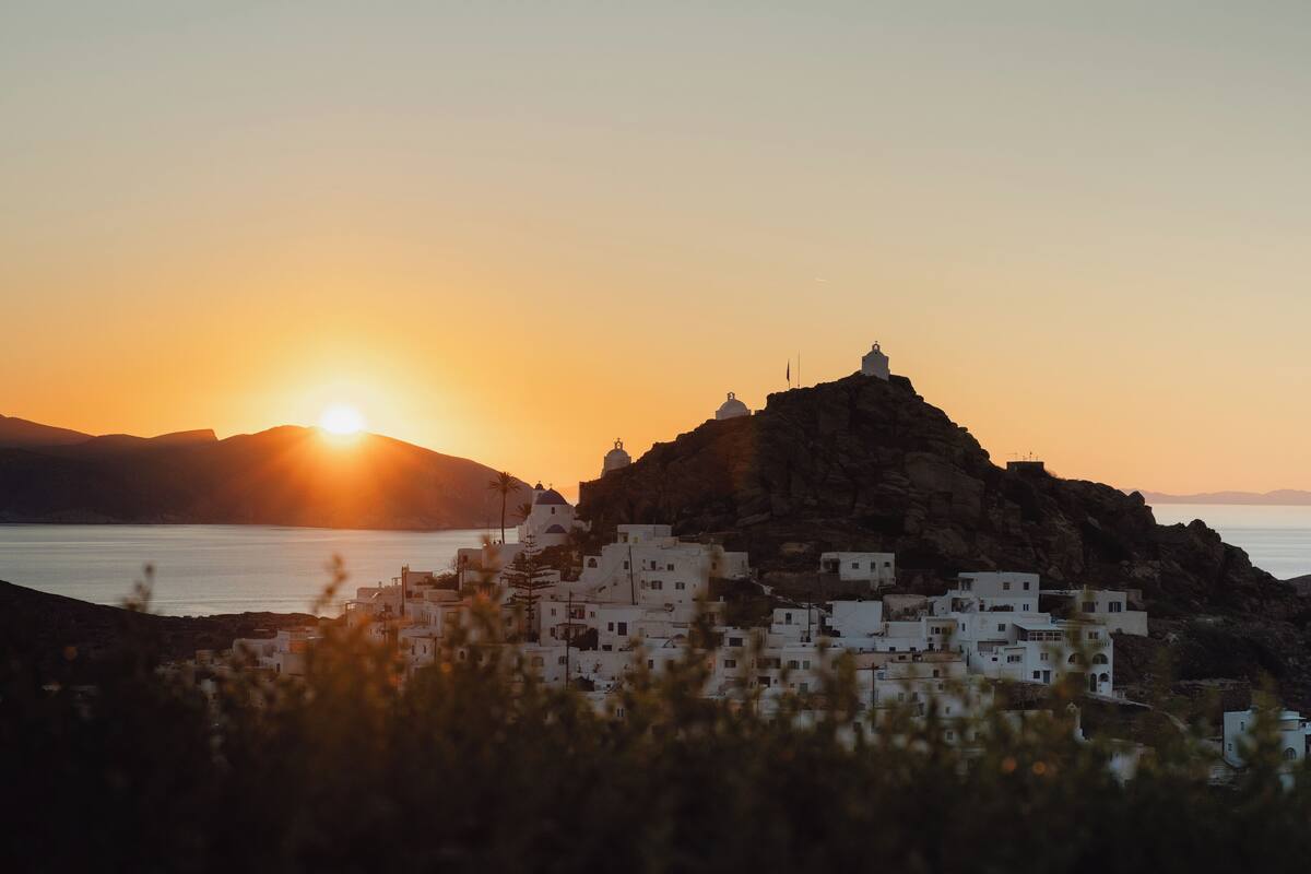 A coastal village with white buildings, a prominent hilltop structure, and the sun setting over the sea.