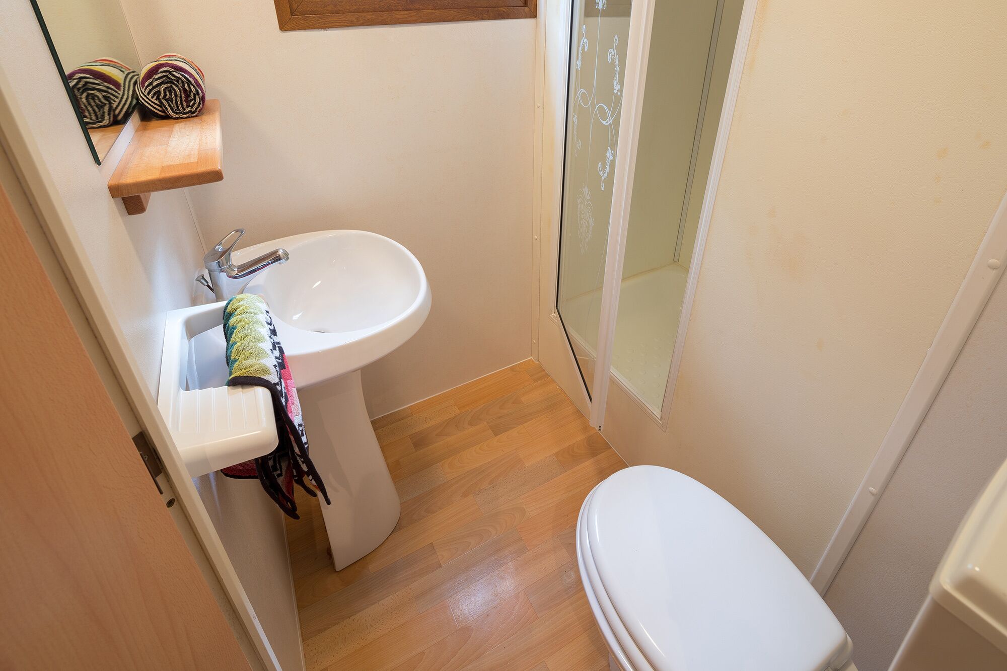 Une petite salle de bain avec un lavabo blanc, des toilettes et une étagère en bois sur laquelle est posée une serviette.