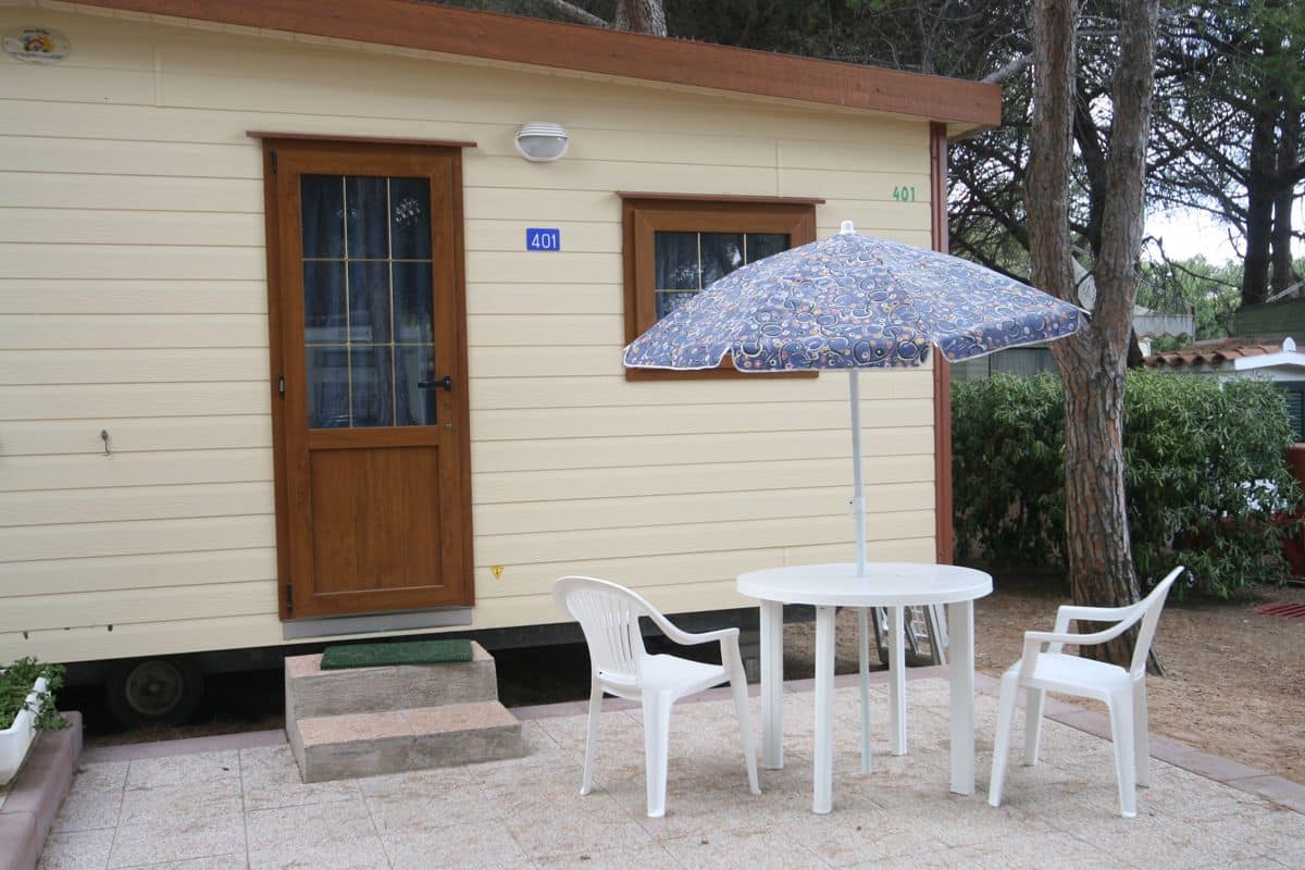 Een laag, enkel verhaal tellend gebouw met een beige gevel, een houten deur en een raam. Op de muur staat het nummer '401', er is een witte paraplu en twee witte plastic stoelen rond een klein tafeltje.