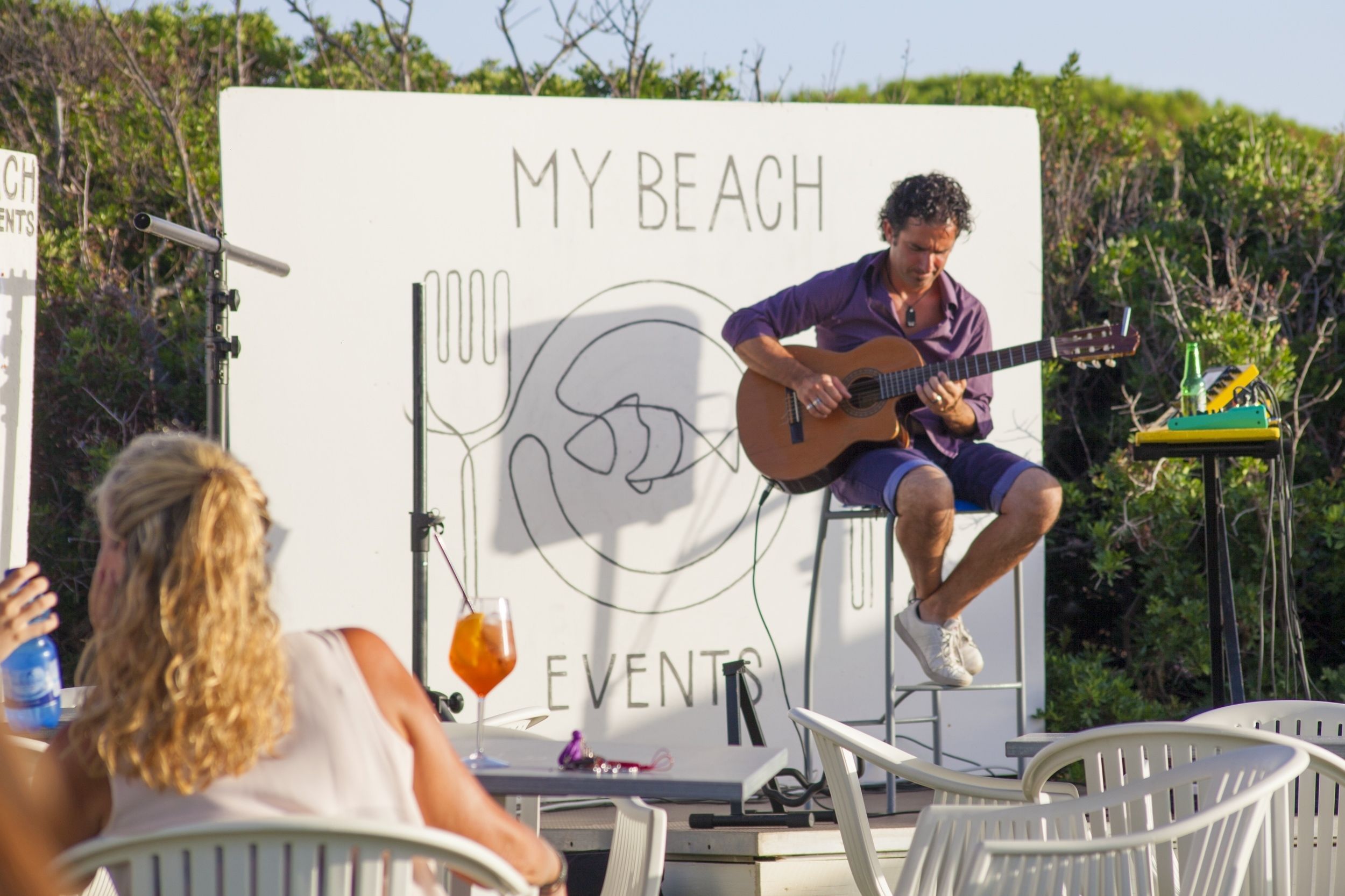 Ein Mann spielt Gitarre auf einer Bühne, auf einem Schild steht 'MY BEACH EVENTS'.
