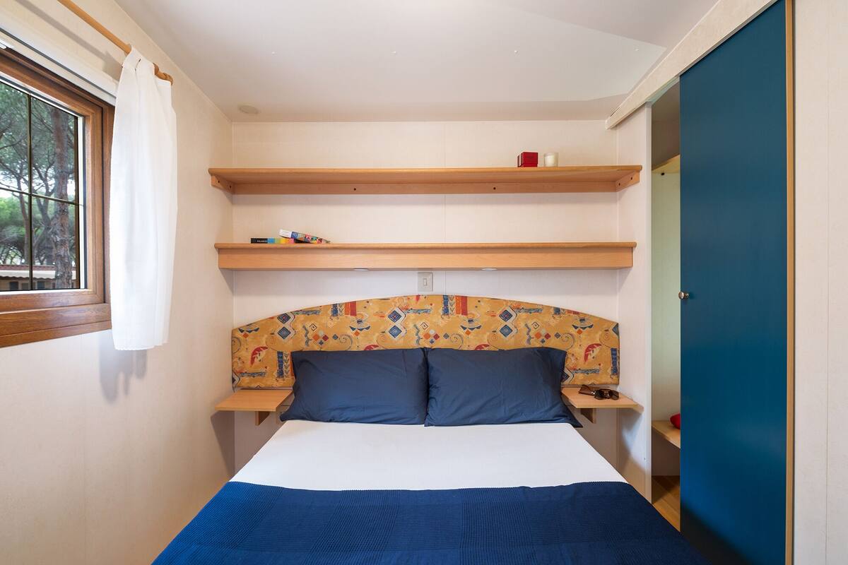 Een slaapkamer met een bed, twee nachtkastjes, een raam met gordijnen en ingebouwde planken met spullen erop.