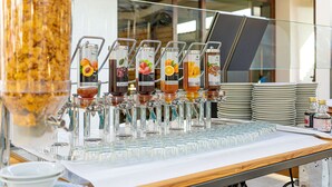 A beverage station with multiple dispensers, a large container of granola, and stacked plates. - Camping Village Baia Blu La Tortuga (Aglientu)
