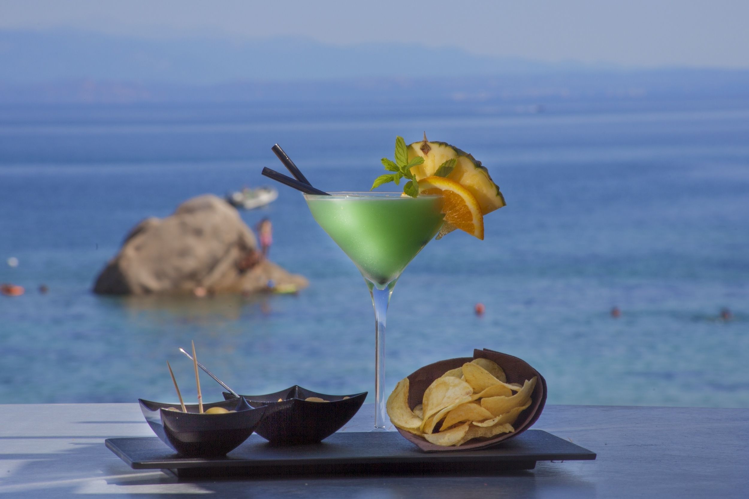 Een groene cocktail met een ananas garnering en een sinaasappelschijfje, een schaal chips en een klein schaaltje met stokjes op een tafel aan zee.