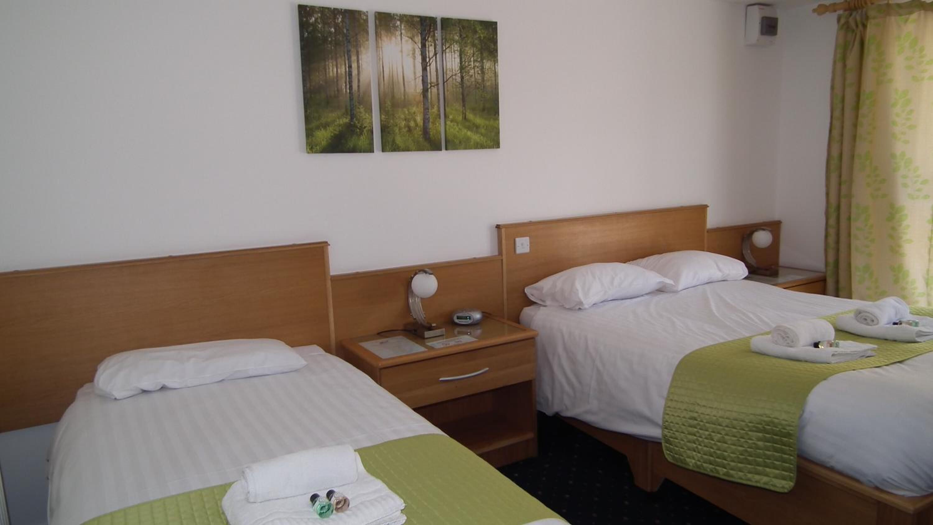 Une chambre d’hôtel avec deux lits, chacun doté d’une tête de lit en bois, de draps blancs et d’une couette verte. On y trouve des tables de chevet avec des lampes et une horloge.