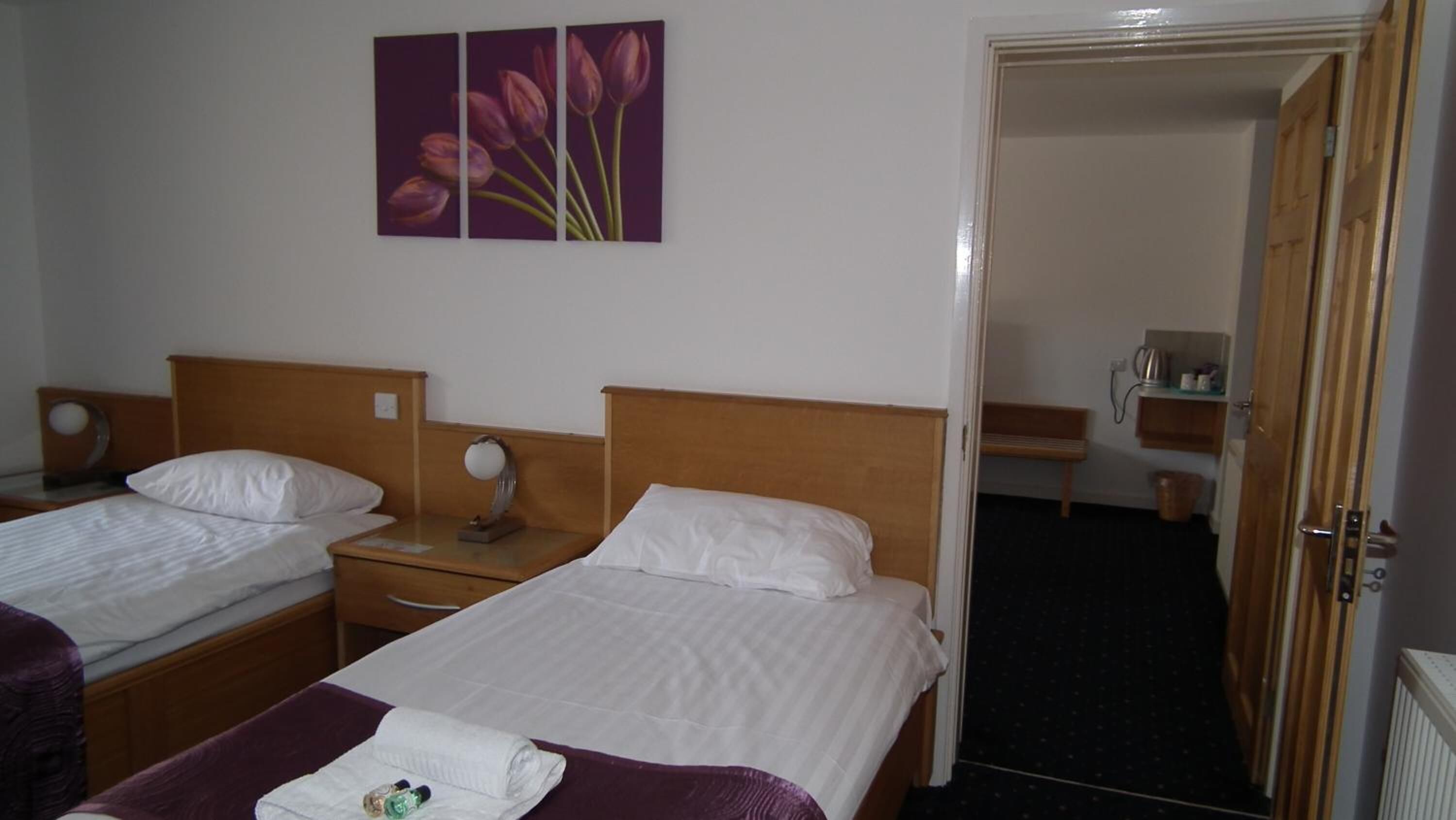 A hotel room with two single beds, a wooden headboard, a bedside table with a lamp, a small mirror, and a painting of flowers on the wall.