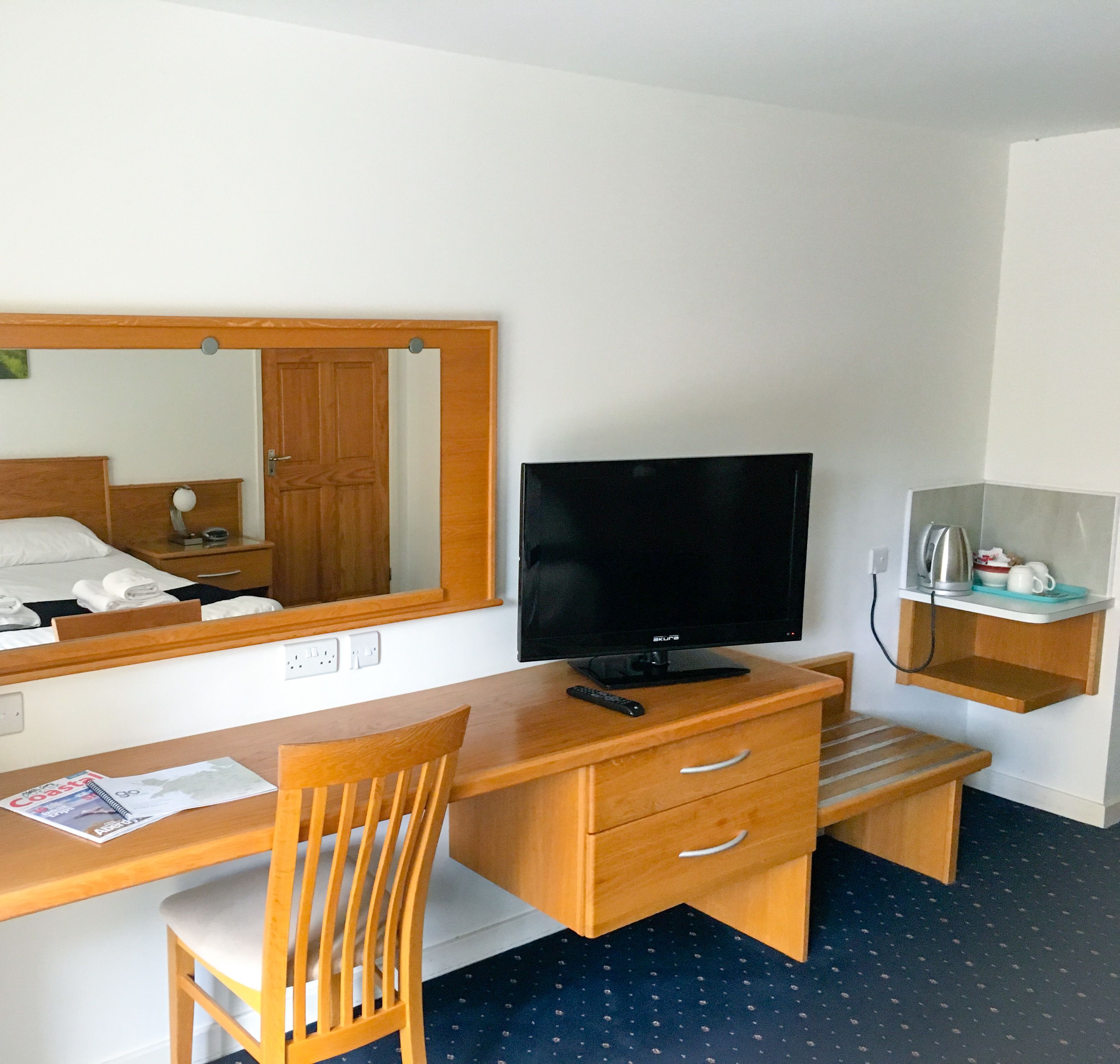 A hotel room with a wooden desk, a television, a chair, a mirror, and a small shelf with a kettle and cups.
