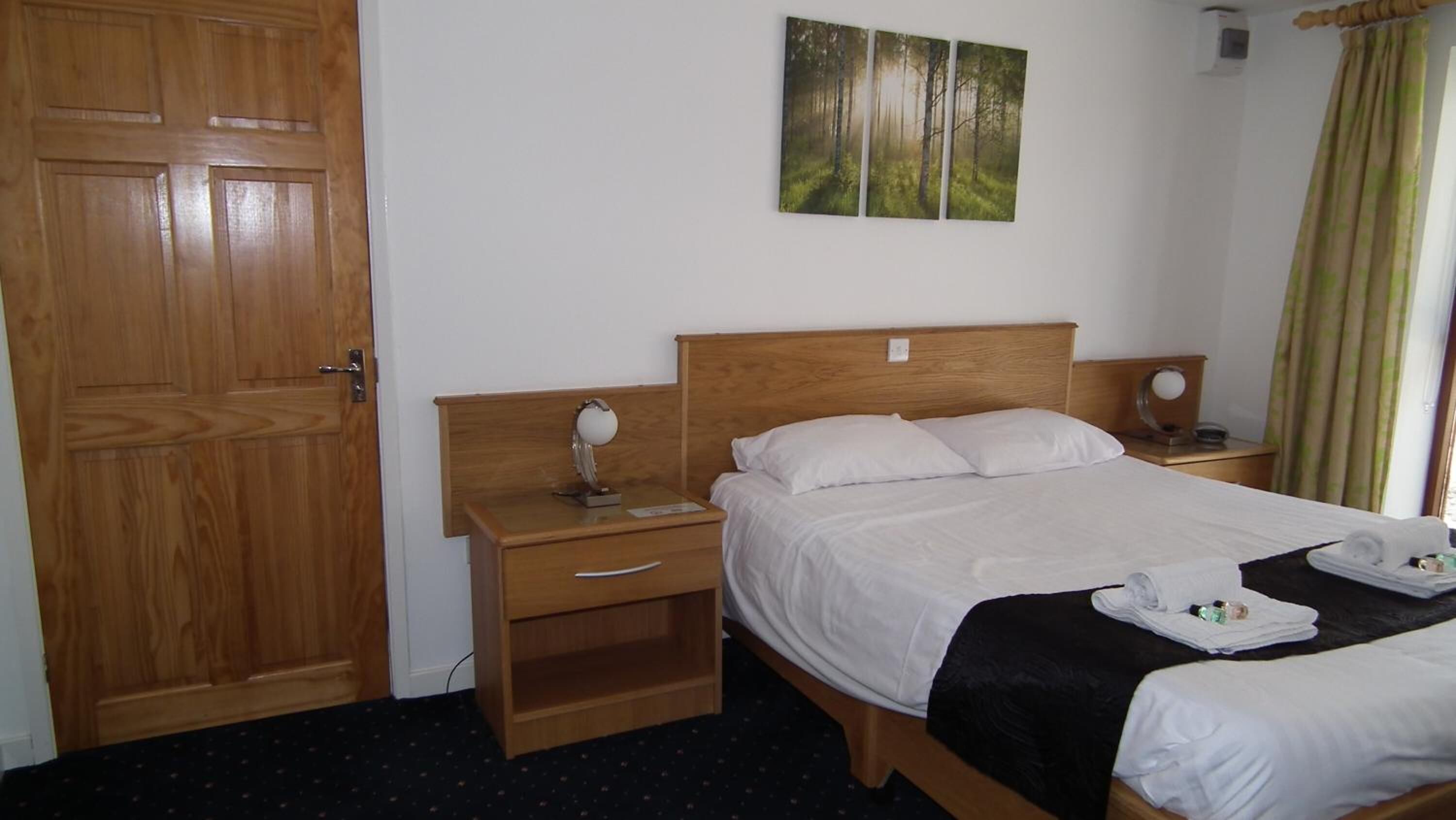 A hotel room with a wooden bed, a nightstand with a lamp, a small shelf, and a painting on the wall.