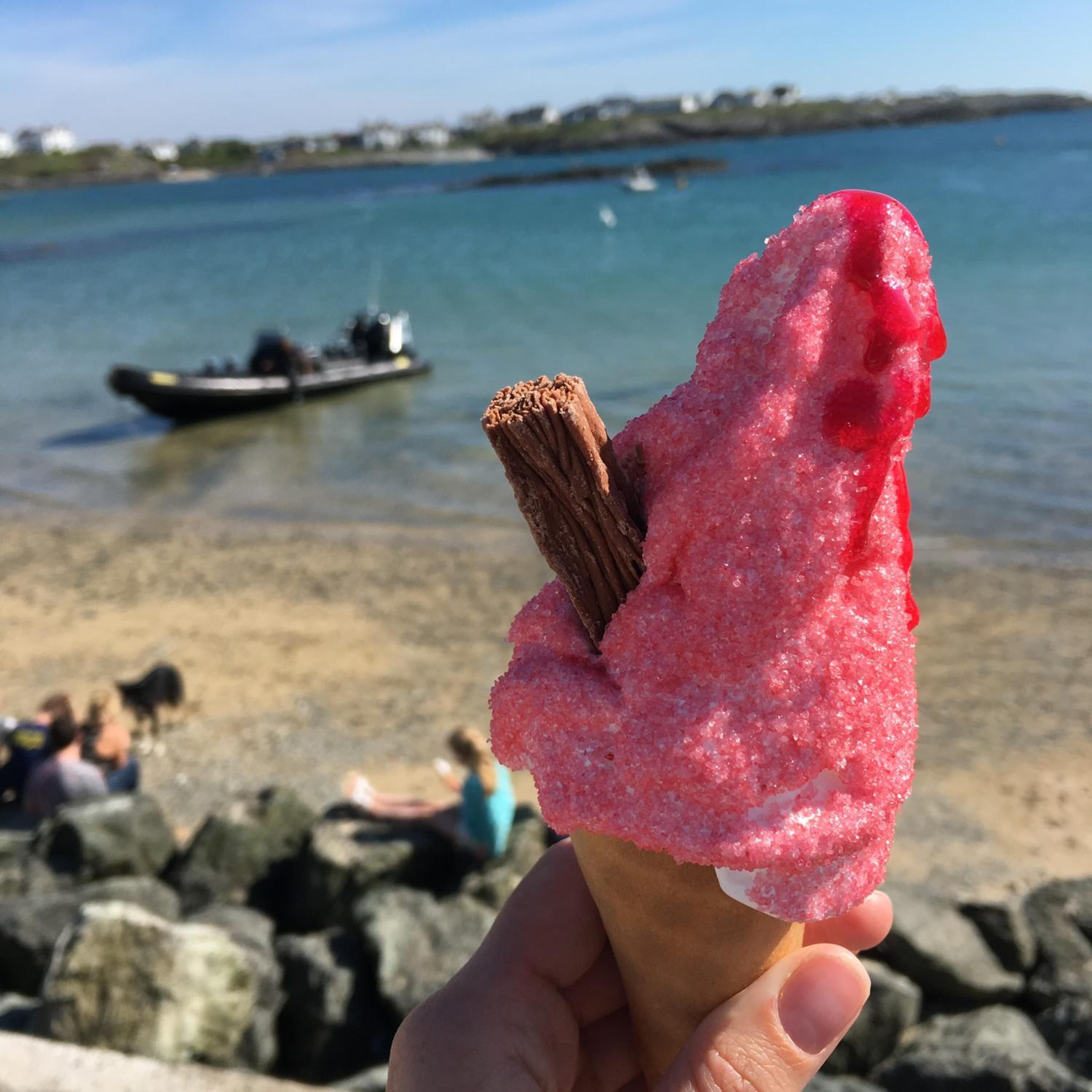Una mano sostiene un cono de helado rosa con palito de chocolate, en un entorno costero con un barco al fondo.