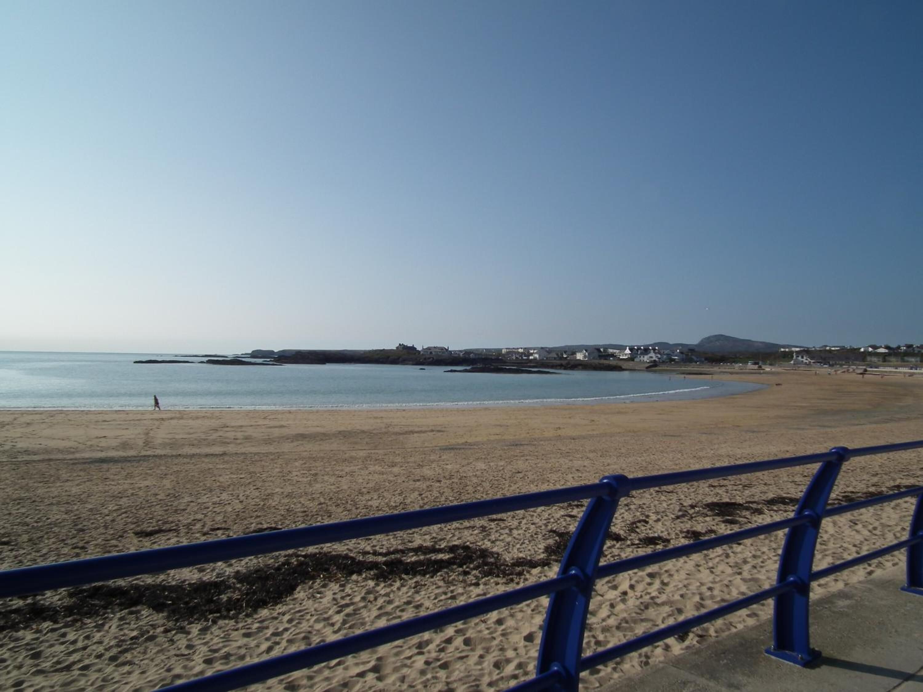 A beach with a blue railing in the foreground, a clear sky, and buildings in the background.