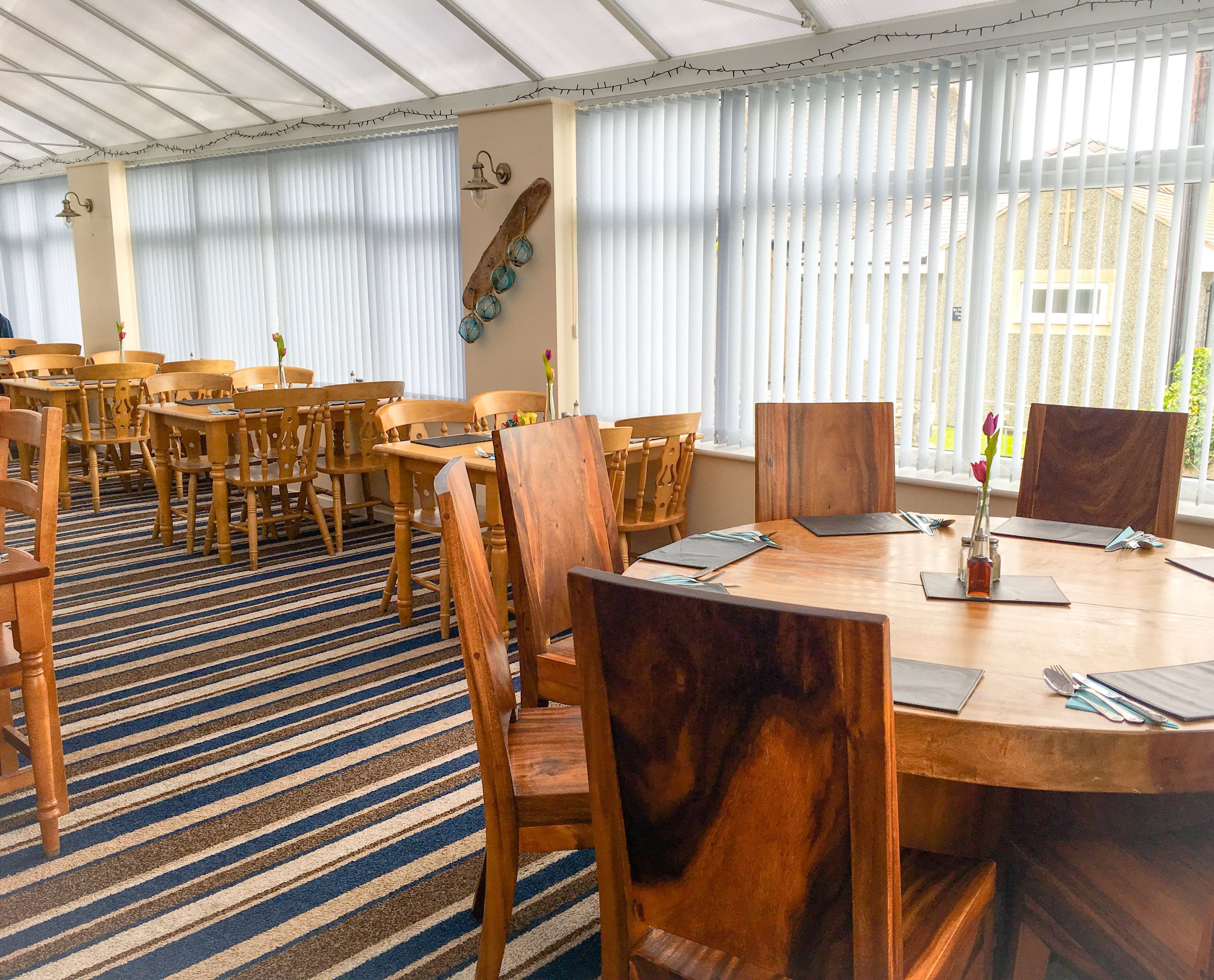 A dining area with wooden tables and chairs, a striped carpet, and a large window with blinds.