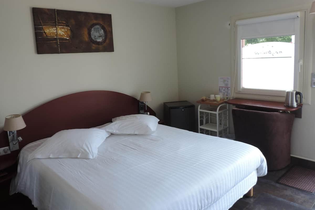 A hotel room with a bed, a desk with a kettle, a small cabinet, and a window.
