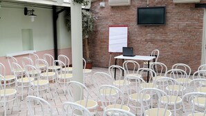 Un área de estar al aire libre con techo, con sillas y mesas blancas, un muro de ladrillo y un televisor montado.