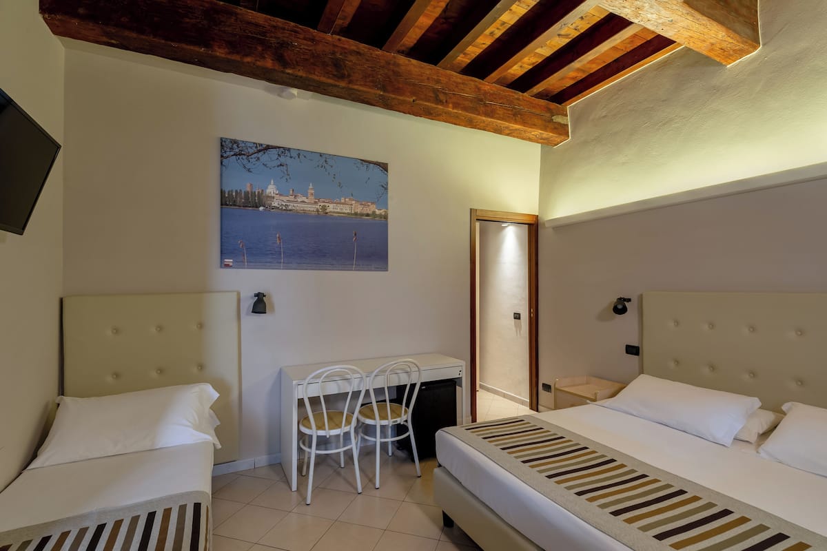 Ein Hotelzimmer mit zwei Betten, einem Schreibtisch mit Stühlen, einem Stadtbildgemälde und einem Holzbalken an der Decke.