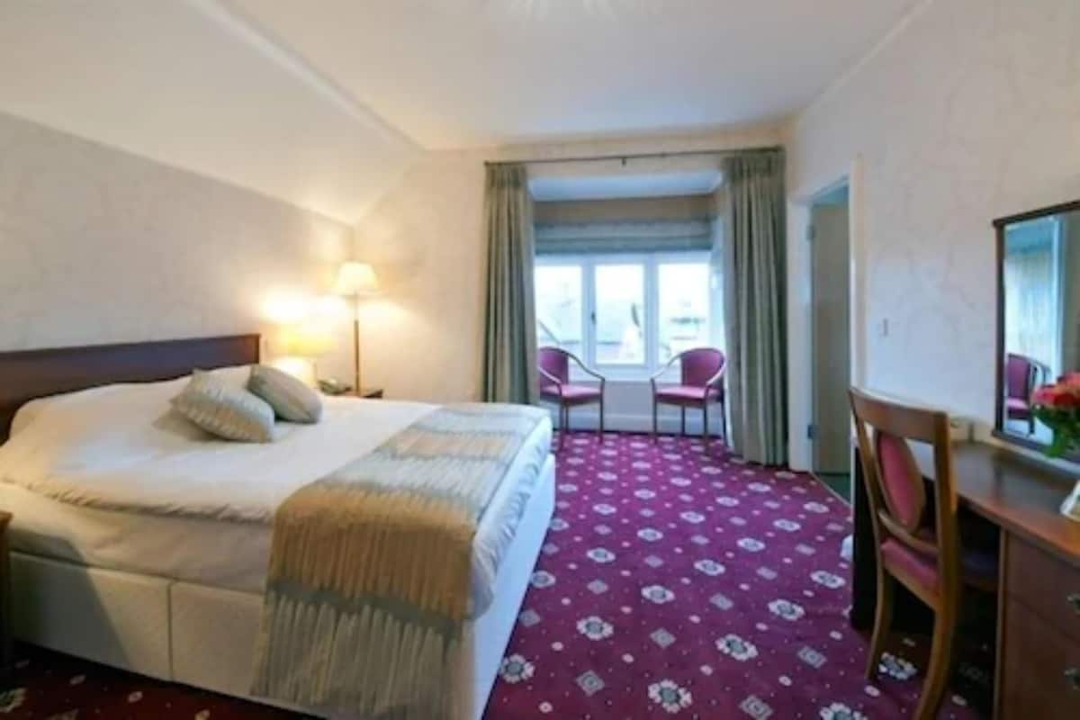 Une chambre d’hôtel comprenant un lit, un bureau, deux chaises, une fenêtre avec des rideaux et une moquette à motifs.