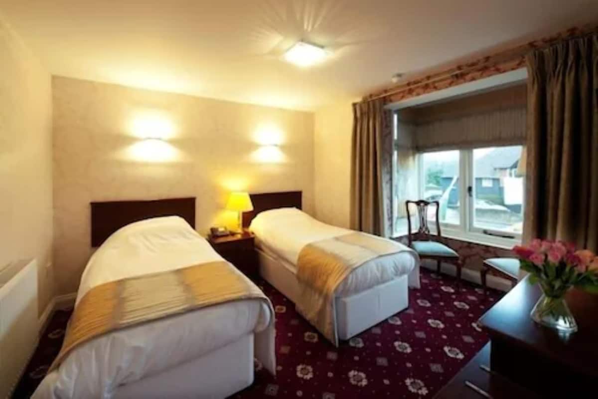 Une chambre d’hôtel avec deux lits simples, un petit bureau avec une chaise, une fenêtre avec des rideaux et une lampe sur la table de chevet.