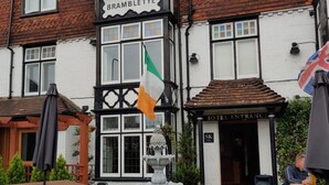 Een traditionele pub met een bord waarop 'BRAMBLETT' staat, terras met tafels en stoelen, een fontein en een vlag.