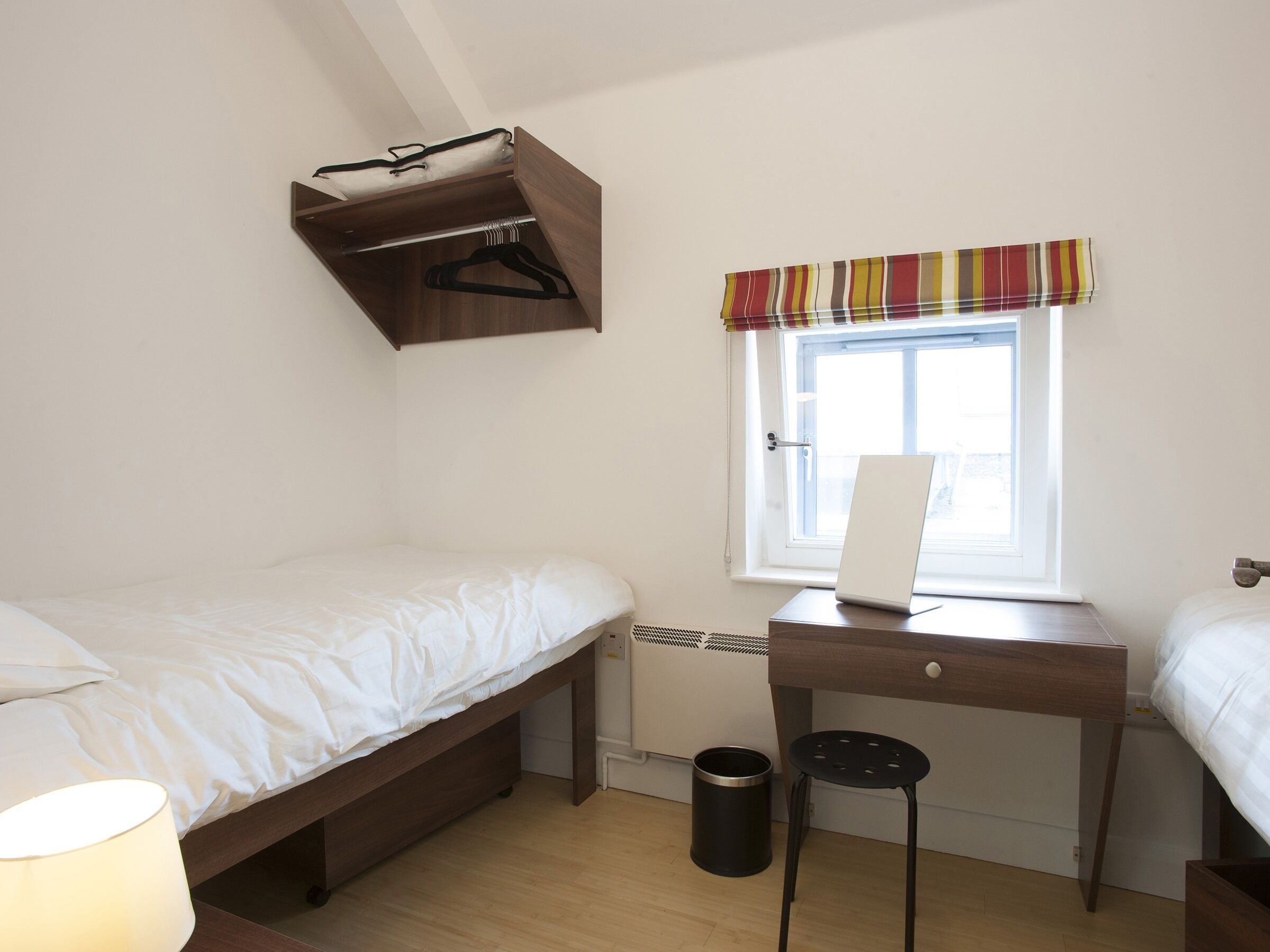 Un dormitorio con cama, un escritorio con computadora portátil, un estante colgante, una ventana con cortinas y un cesto de basura.