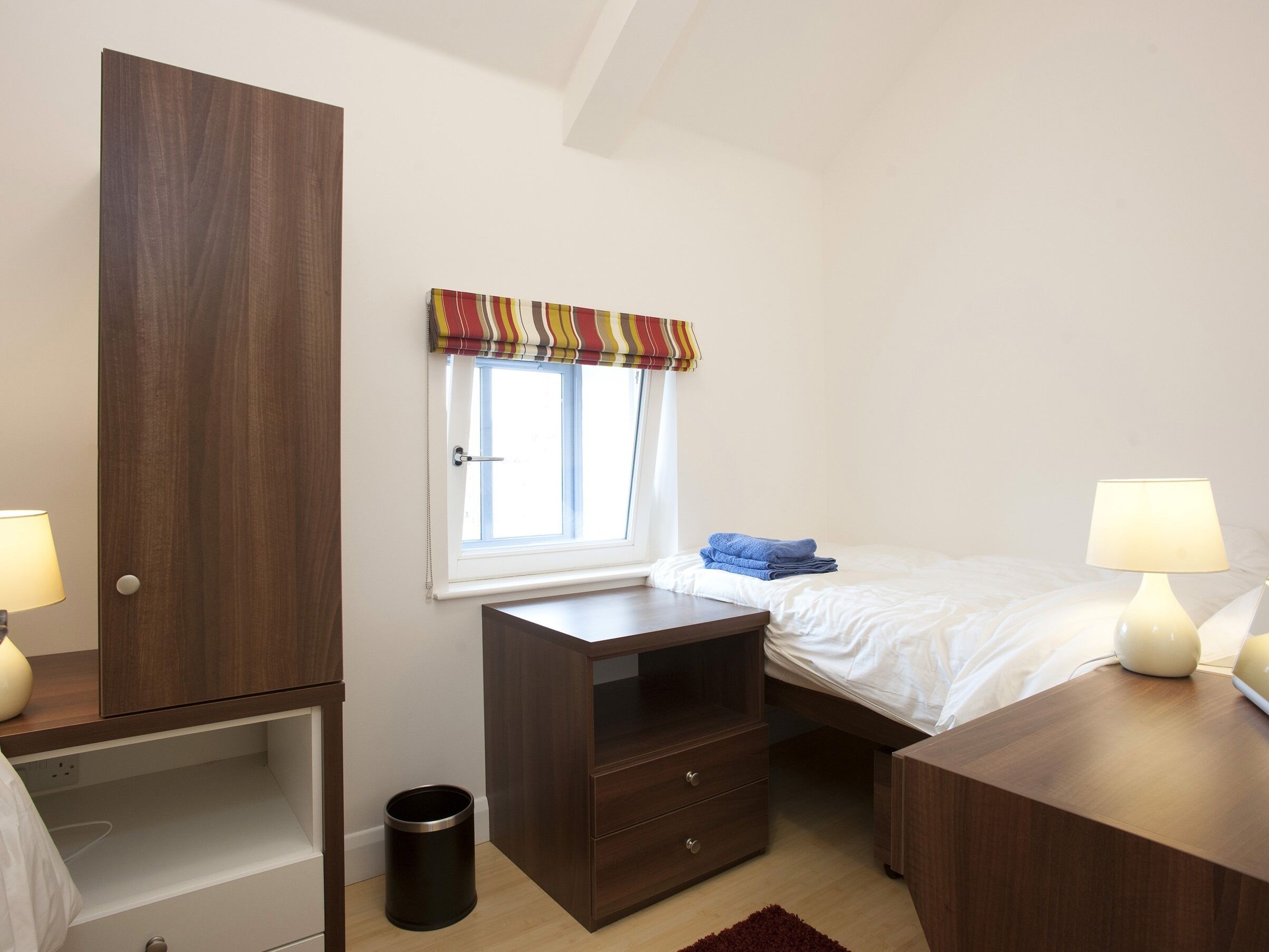 Een slaapkamer met een bed, nachtkastjes, een kledingkast, een raam met gordijnen en een kleine prullenbak.