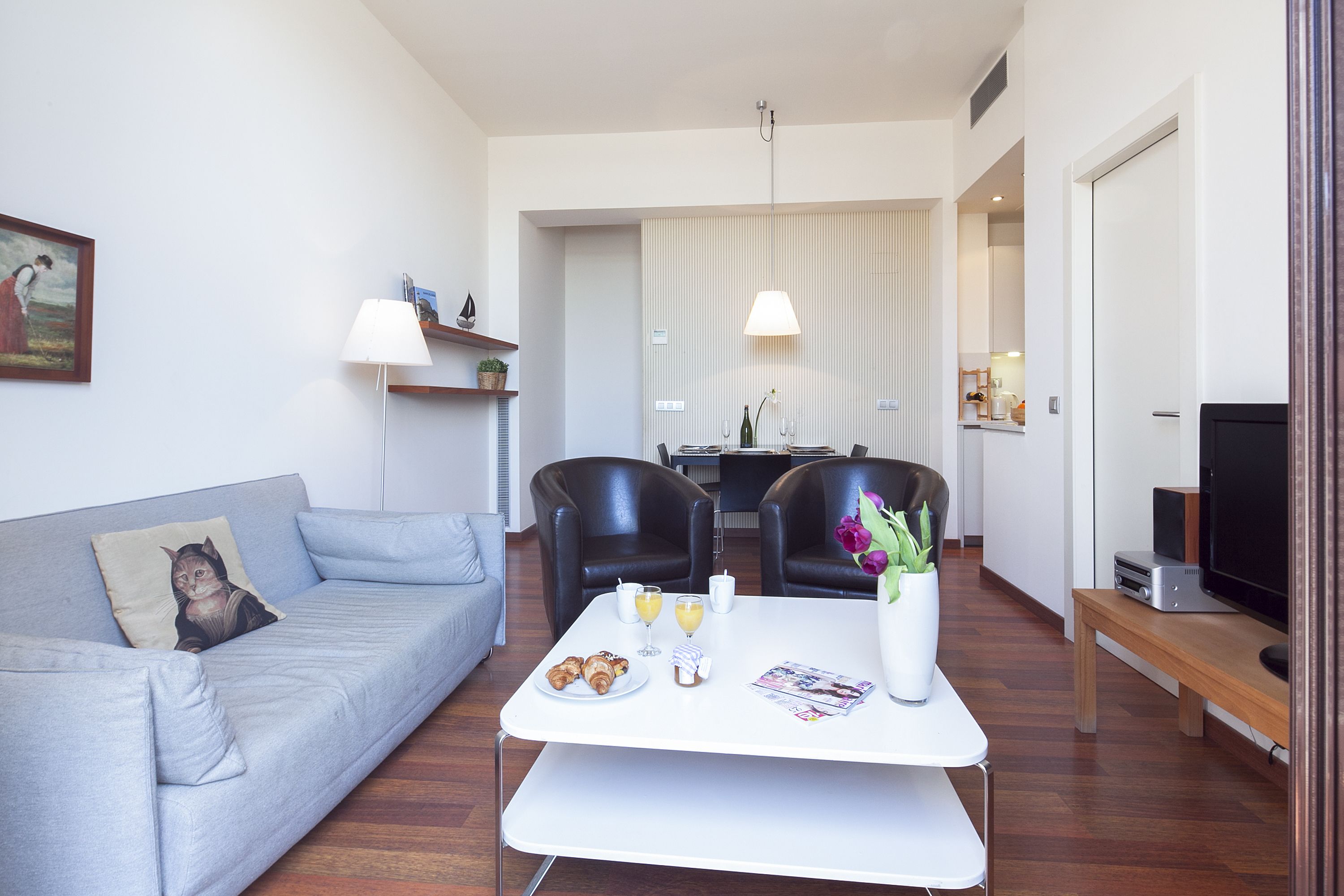 Una sala de estar moderna con un sofá gris, una mesa de centro blanca con alimentos para el desayuno, un televisor y una zona de comedor con sillas y una mesa.